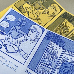 Two comic zines open on pages showing comic panels of a girl sitting at a desk drawing in a sketchbook, and a girl opening a fridge.