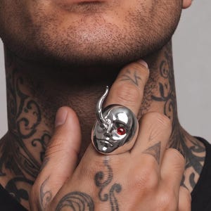 May include: A polished silver ring featuring a stylized face with a single red gemstone eye and a curved horn-like protrusion extending from the forehead. The ring has a smooth, reflective metallic finish and covers the finger with a bold design.