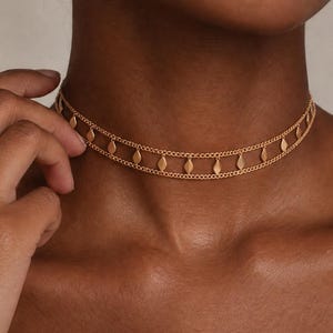 Op de afbeelding: Een goudkleurige choker-ketting bestaande uit twee fijne parallelle kettingen verbonden door kleine, gelijkmatig verdeelde ruitvormige bedels. Het sieraad sluit nauw aan op de hals en heeft een metalen afwerking en een gestructureerd ontwerp.