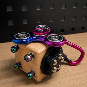 Könnte beinhalten: Ein hölzernes Fidget-Würfel-Spielzeug mit einem mehrfarbigen Metall-Spinner auf der Oberseite, vier schillernden Schrauben an der Seite und einem schwarzen runden Bauteil mit silbernen Kugellagern. Ein pinker Karabiner mit dem Text Werkstater ist seitlich befestigt.