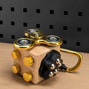 Może przedstawiać: Drewniana kostka typu fidget z czterema złotymi, teksturowanymi pokrętłami z jednej strony, czarnym plastikowym panelem z pięcioma srebrnymi metalowymi łożyskami kulkowymi, złotym metalowym spinnerem na górze i złotym karabińczykiem z boku.