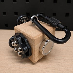 Könnte beinhalten: Ein hölzerner Fidget-Würfel mit einem schwarzen Metallkarabiner an der Oberseite. Eine Seite zeigt ein schwarzes Kunststoffelement mit vier silbernen Kugellagern, während eine andere Seite eine runde silberne Metallscheibe aufweist.