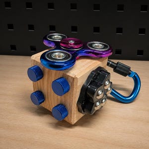 Könnte beinhalten: Ein hölzerner Fidget-Würfel mit einem mehrfarbigen metallischen Spinner auf der Oberseite, vier blauen, strukturierten Knöpfen an der Seite, einem schwarzen Kunststoffelement mit fünf silbernen Metallkugeln und einem blauen Karabiner an der Seite.