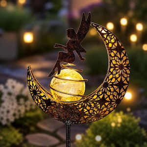 May include: A metal garden stake featuring a crescent moon with an intricate cutout pattern and a dark bronze finish. A silhouette of a fairy sits atop a glowing, crackled glass orb held within a spiral wire frame centered in the moon.