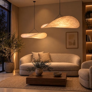 May include: Two large, wide, oval-shaped pendant lights with a textured, beige, paper-like material suspended from the ceiling by thin black wires. The lights emit a warm, soft glow, illuminating the living room space below.