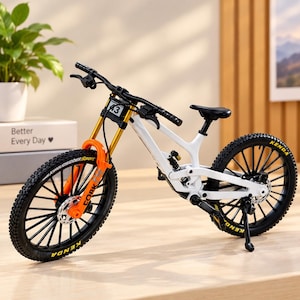 May include: A miniature white mountain bike model featuring a bright orange front fork, black handlebars, and black tires with Kenda branding. The frame includes a small number plate with the text 43, a rear suspension component, and a kickstand.