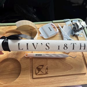 Op de afbeelding: Een witte schuimcilinder met de zwarte tekst LIV'S 18TH, vastgebonden met een zwart satijnen lint. De cilinder ligt boven een houten plank met een kleine paraplu- en flesafbeelding, naast een wit pelmesje en een witte schraper.