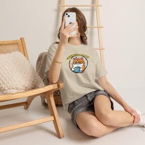 Op de afbeelding: Een beige t-shirt met korte mouwen en een afbeelding van een oranje cyperse kat met een bril die een blauwe mok vasthoudt. De tekst boven de kat luidt CAFFEINATED & CRANKY in gele letters, omringd door een cirkelvormige rand met gekleurde stippen.