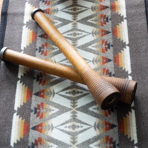 May include: Two wooden spools with metal ends rest on a patterned textile. The spools are cylindrical with a ridged design on one end. The textile features a geometric pattern in shades of brown, white, orange, and yellow.