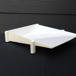 May include: A white square tray with a sloped design, featuring four cylindrical legs at each corner. The tray has a smooth, solid white finish and an open top, with the sides gradually increasing in height toward the back corners.