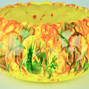 May include: A yellow ceramic bowl with a textured, pineapple-like design. The bowl features a vibrant color scheme of yellow, orange, and green, with red accents. The rim has a scalloped edge.
