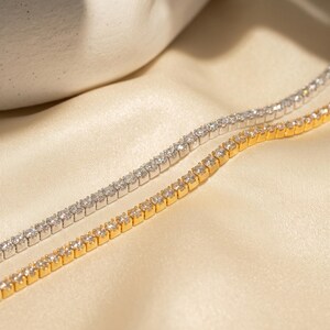 May include: Two tennis bracelets, one in a silver-toned metal and one in a gold-toned metal, each featuring a continuous row of small, clear, round-cut stones set in a prong setting, arranged side-by-side in a parallel, linear formation.