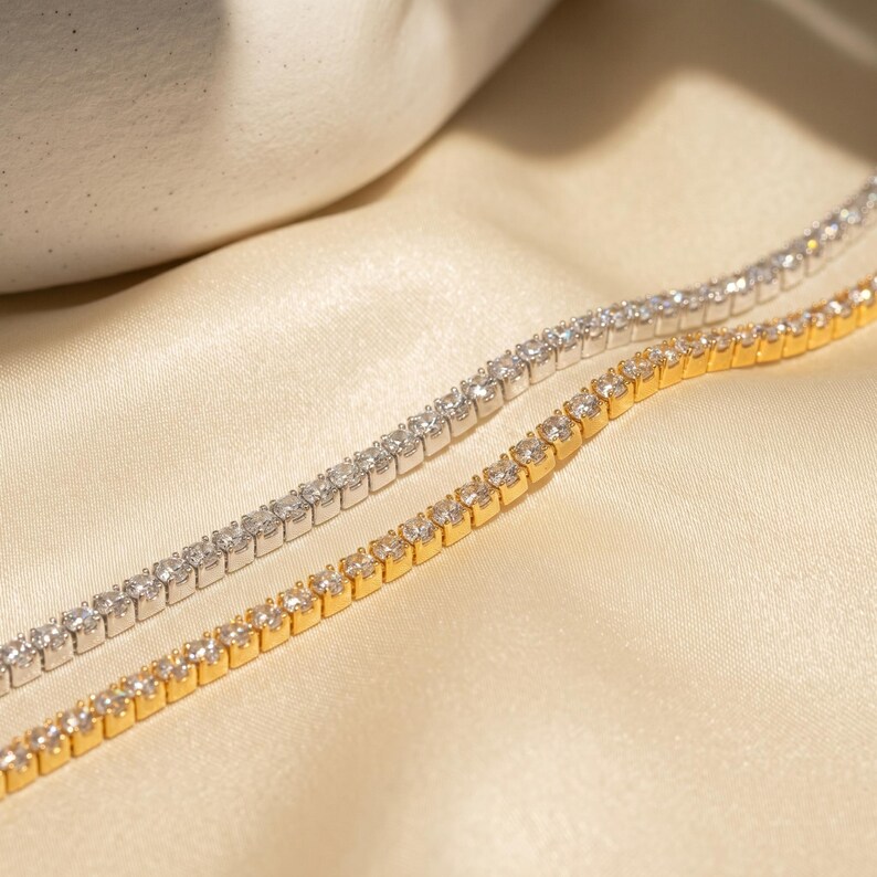 May include: Two tennis bracelets, one in a silver-toned metal and one in a gold-toned metal, each featuring a continuous row of small, clear, round-cut stones set in a prong setting, arranged side-by-side in a parallel, linear formation.