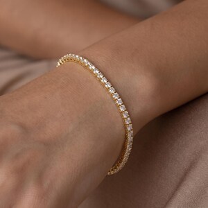 May include: A gold tennis bracelet featuring a continuous row of round, clear, sparkling stones set in a polished metal frame. The bracelet encircles the wrist, showcasing a uniform design with each stone securely held in a prong setting.