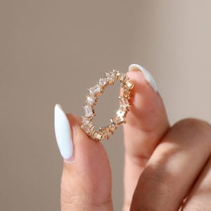 Peut inclure: Une alliance en or composée d'une rangée continue de pierres transparentes alternant entre une taille émeraude et une taille ronde, serties dans une monture en métal poli. La bague présente un motif répétitif de gemmes rectangulaires et rondes.