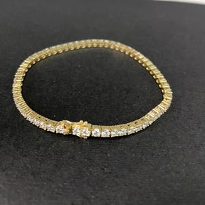 May include: A gold-toned tennis bracelet featuring a continuous row of round, clear, faceted stones set in a metal frame. The bracelet includes a secure box clasp mechanism with a safety latch, creating a flexible circular band design.