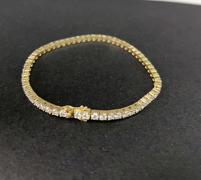 May include: A gold-toned tennis bracelet featuring a continuous row of round, clear, faceted stones set in a metal frame. The bracelet includes a secure box clasp mechanism with a safety latch, creating a flexible circular band design.