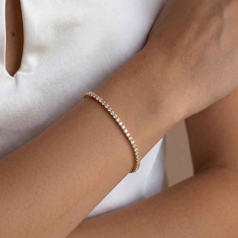 May include: A gold-toned tennis bracelet featuring a continuous row of small, clear, round-cut stones set in a metallic band. The bracelet is worn around the wrist, showcasing a flexible design with a uniform arrangement of sparkling, reflective gems.
