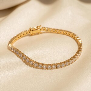 May include: A gold-toned tennis bracelet featuring a continuous row of clear, round-cut stones set in a metallic frame. The bracelet includes a secure box clasp mechanism at one end, designed to hold the circular chain together when worn.
