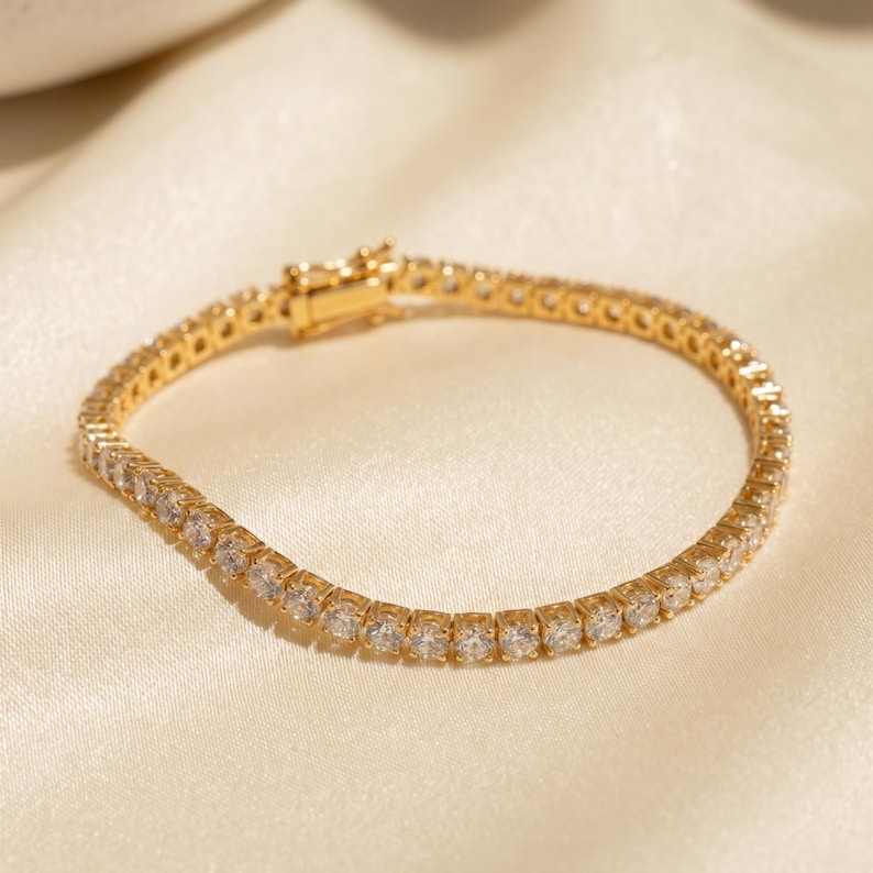 May include: A gold-toned tennis bracelet featuring a continuous row of clear, round-cut stones set in a metallic frame. The bracelet includes a secure box clasp mechanism at one end, designed to hold the circular chain together when worn.