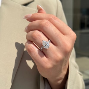 Puede incluir: Un anillo de tono plateado con una gran piedra central de corte en forma de pera y una banda acentuada con pequeñas piedras engastadas en pavé. El anillo se lleva en un dedo, mostrando la claridad facetada de la gema central.