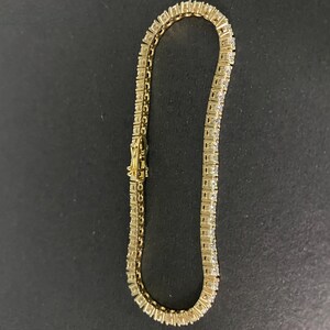 May include: A gold-toned tennis bracelet featuring a continuous row of square-cut clear stones set in a metal frame. The bracelet includes a secure box clasp mechanism with a safety latch, showcasing a uniform link design throughout the piece.