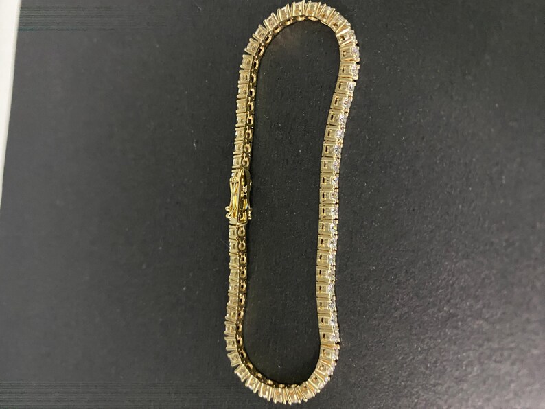 May include: A gold-toned tennis bracelet featuring a continuous row of square-cut clear stones set in a metal frame. The bracelet includes a secure box clasp mechanism with a safety latch, showcasing a uniform link design throughout the piece.