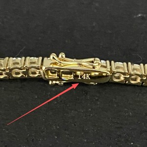 May include: A gold-toned tennis bracelet featuring a series of clear, round-cut stones set in a metal chain. The clasp mechanism is clearly visible and features the engraved text 14K, with a thin red arrow pointing directly to the marking.