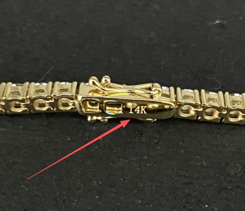 May include: A gold-toned tennis bracelet featuring a series of clear, round-cut stones set in a metal chain. The clasp mechanism is clearly visible and features the engraved text 14K, with a thin red arrow pointing directly to the marking.