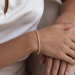 May include: A gold tennis bracelet featuring a continuous row of small, round, clear stones set in a metallic band. The bracelet encircles the wrist, showcasing a uniform design with reflective surfaces that catch the light, creating a sparkling effect.