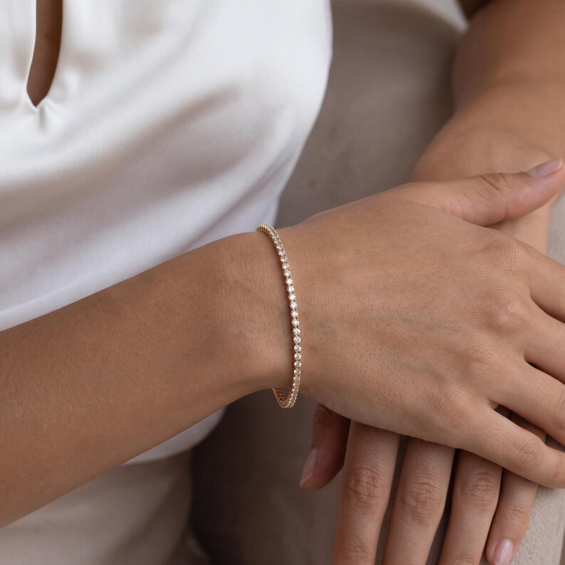 May include: A gold tennis bracelet featuring a continuous row of small, round, clear stones set in a metallic band. The bracelet encircles the wrist, showcasing a uniform design with reflective surfaces that catch the light, creating a sparkling effect.