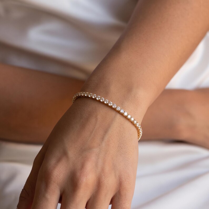 May include: A gold-toned tennis bracelet featuring a continuous row of small, clear, round-cut stones set in a metallic frame. The bracelet encircles the wrist, showcasing a uniform design with reflective surfaces and a classic, flexible link structure.