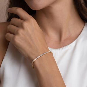 May include: A gold-toned tennis bracelet featuring a continuous row of small, clear, round-cut stones set in a metallic band. The bracelet encircles the wrist, showcasing a uniform design with a polished finish and sparkling, reflective facets.