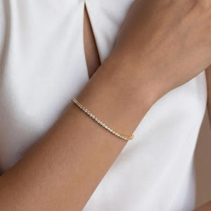 May include: A gold-toned tennis bracelet featuring a continuous row of small, clear, round-cut stones set in a metallic band. The bracelet is worn around the wrist, showcasing a uniform design with reflective surfaces and a polished finish.