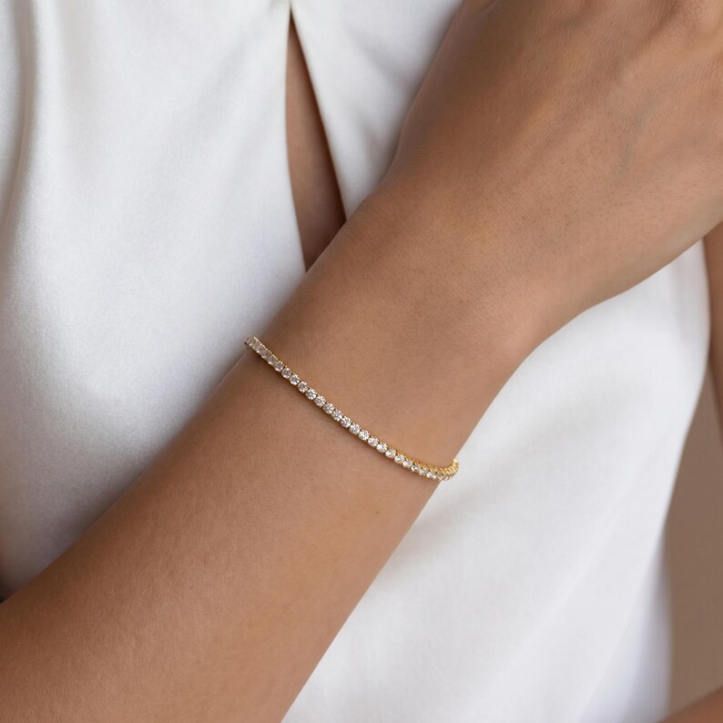 May include: A gold-toned tennis bracelet featuring a continuous row of small, clear, round-cut stones set in a metallic band. The bracelet is worn around the wrist, showcasing a uniform design with reflective surfaces and a polished finish.