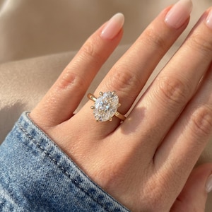 Puede incluir: Un anillo de oro con una gran piedra preciosa transparente de talla ovalada, montada en un engaste de cuatro garras. La banda es fina y pulida, mostrando un diseño clásico que resalta el brillo y la claridad de la piedra ovalada central.