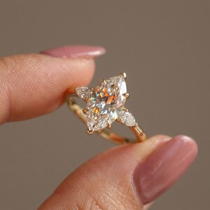 Puede incluir: Un anillo de oro con una gran piedra central de corte marquesa flanqueada por dos piedras laterales más pequeñas de corte pera. La banda es fina y pulida, sosteniendo las tres gemas claras y facetadas en un engaste de garras.