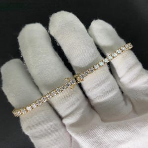 May include: A gold-toned tennis bracelet featuring a continuous row of round, clear, faceted stones set in a metal frame. The bracelet includes a secure box clasp mechanism with a safety latch, showcasing a uniform design of sparkling stones.