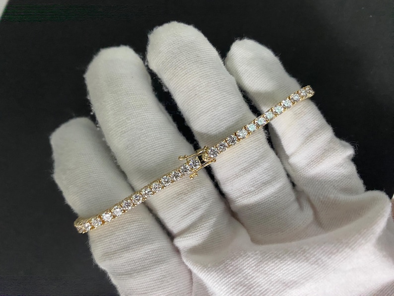 May include: A gold-toned tennis bracelet featuring a continuous row of round, clear, faceted stones set in a metal frame. The bracelet includes a secure box clasp mechanism with a safety latch, showcasing a uniform design of sparkling stones.