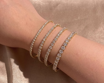 May include: A set of four gold-tone tennis bracelets, each featuring a continuous row of round, clear, faceted stones set in a metallic frame. The bracelets are worn together, displaying a uniform design with sparkling, reflective surfaces.