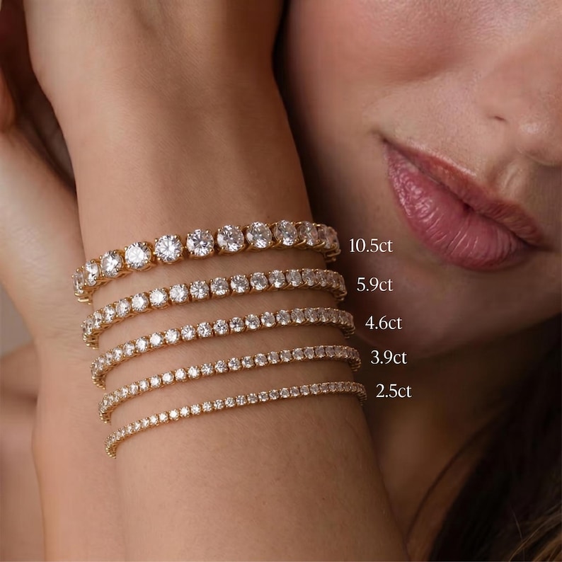 May include: Five gold tennis bracelets featuring round-cut clear stones in varying sizes, labeled with carat weights 10.5ct, 5.9ct, 4.6ct, 3.9ct, and 2.5ct. The bracelets are arranged in a stack, showcasing different stone diameters and metal settings.