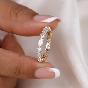 May include: A gold band ring featuring an alternating pattern of clear baguette-cut and round-cut stones set in a continuous row. The metal band has a polished finish, and the stones are arranged to create a repeating sequence around the entire ring.