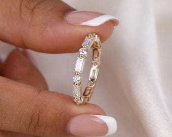 May include: A gold band ring featuring an alternating pattern of clear baguette-cut and round-cut stones set in a continuous row. The metal band has a polished finish, and the stones are arranged to create a repeating sequence around the entire ring.