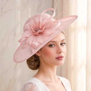 May include: A wide-brimmed dusty pink fascinator hat featuring a large, layered fabric flower accent on the side. The hat is adorned with delicate, thin, curved wire loops that extend upwards from the centre of the floral arrangement.
