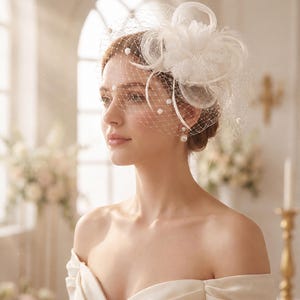 May include: A white fascinator headpiece featuring a delicate netting veil adorned with small white pearls. The headpiece is decorated with a layered fabric flower, looped ribbon accents, and thin, wispy feathers, creating a textured accessory.