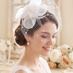 May include: A white fascinator headpiece featuring a structured fabric flower, delicate feathers, and a birdcage veil adorned with small white polka dots. The accessory is secured to the hair, providing a decorative accent with a classic netting design.