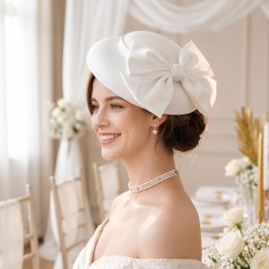 May include: A white pillbox hat featuring a large, structured bow on the side. The hat has a smooth, matte finish and is worn with a multi-strand pearl choker necklace and a single pearl drop earring, creating a coordinated and formal appearance.