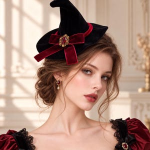 May include: A black velvet witch hat featuring a deep red velvet ribbon bow and a central gold-toned brooch with a red gemstone. The hat is adorned with a matching red velvet band around the base of the crown, creating a cohesive and dark aesthetic.
