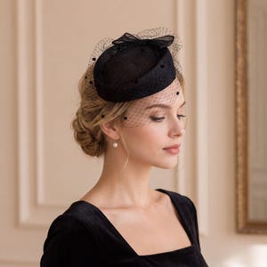 May include: A black pillbox hat featuring a structured circular crown and a delicate netting veil. The veil is adorned with small, evenly spaced black dots and is secured at the top with a small, matching black fabric bow detail.