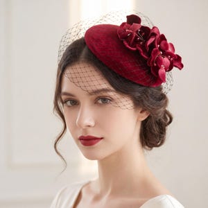 Könnte beinhalten: Ein tiefroter Fascinator aus Samt mit einer kreisförmigen Basis, die mit einer Gruppe passender Stoffblumen und kleinen Perlen verziert ist. Ein schwarzer Netzschleier erstreckt sich vom Hut und bedeckt den oberen Teil des Gesichts mit einem feinen Muster.